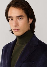 Blazer de pana azul marino sobre un cuello de tortuga verde oscuro. El blazer tiene solapas con muescas y un bolsillo, textura suave y corte estructurado.
