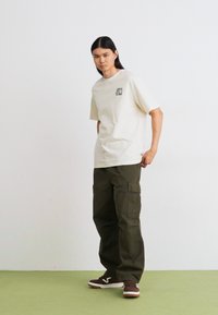 Beige cotton t-shirt with a front graphic pocket and olive cargo pants. Textured fabric, relaxed fit, paired with brown sneakers.
