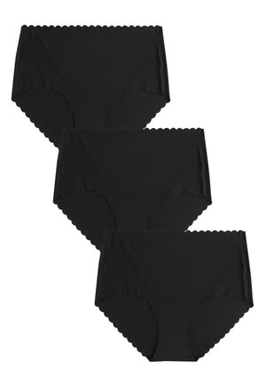 Žena nosi čierne bezšvové spodné prádlo s vlnkovaným okrajom a zladený dlhý rukáv, stojaca na jednofarebnom pozadí.