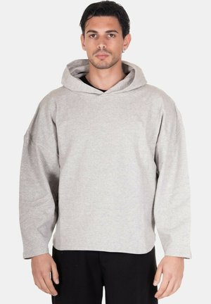 Giovane uomo con capelli scuri che indossa una felpa oversize grigio chiaro e pantaloni neri, in piedi davanti a uno sfondo bianco semplice.