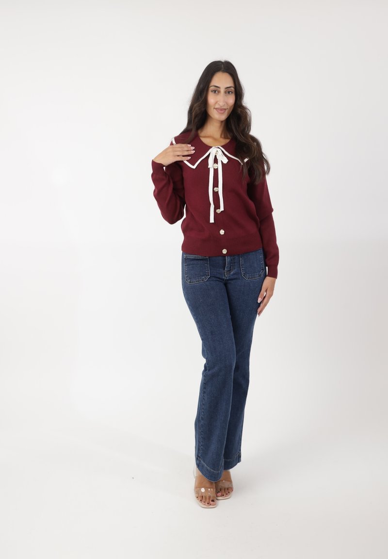 Suéter de punto burdeos con pajarita blanca, con botones en el frente, combinado con jeans de campana azul oscuro y tacones transparentes, de pie sobre un fondo blanco.