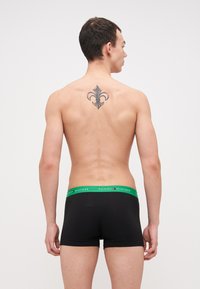 Schwarze Boxershorts mit einem grünen Bund, der das "TOMMY HILFIGER"-Logo trägt. Das Model hat ein Tattoo einer Lilie auf dem Rücken.