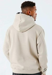 Hoodie en tissu beige clair, doté d'une grande capuche, de poignets côtelés et d'un accent logo sur la manche. Texture lisse avec une coupe décontractée.