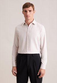 Camicia a maniche lunghe beige chiaro realizzata in un tessuto texturizzato, con colletto a punta e bottoni bianchi, abbinata a pantaloni scuri plissettati.
