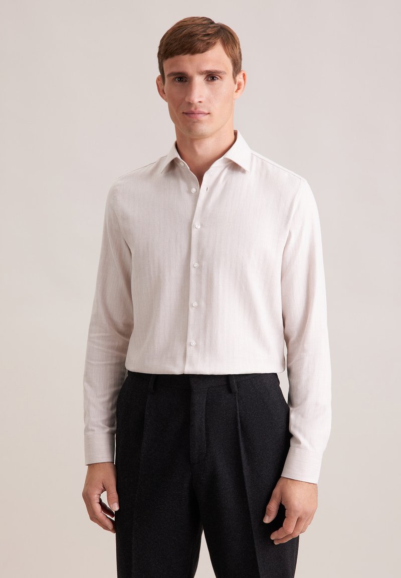 Camicia a maniche lunghe beige chiaro realizzata in un tessuto texturizzato, con colletto a punta e bottoni bianchi, abbinata a pantaloni scuri plissettati.
