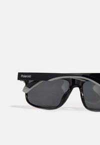 Polaroid napszemüveg fekete műanyag kerettel, szürke kiegészítőkkel, sötét lencsékkel, szögletes kialakítással és merész, lapos felső vonallal.