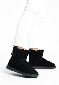 Botas de tobillo de ante negras con un suave forro de piel, que cuentan con un botón decorativo y una correa elástica, suela plana de goma y diseño de punta redonda.