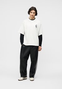 Νεαρό άτομο με μεσαίου μήκους σκούρα μαλλιά φοράει ένα λευκό φαρδύ μπλουζάκι πάνω από ένα μαύρο μακρυμάνικο πουκάμισο, μαύρο φαρδύ παντελόνι και λευκά αθλητικά παπούτσια.