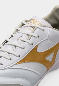 Λευκό αθλητικό παπούτσι με χρυσό λογότυπο και κορδόνια, κοντινή λήψη του επάνω μέρους, που δείχνει το κείμενο της μάρκας και λεπτομερείς ραφές.