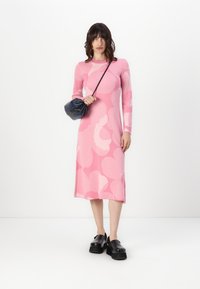 Rochie roz cu mânecă lungă, cu modele abstracte în nuanțe de roz deschis, cu croială dreaptă și lungime midi; asortată cu o geantă mică neagră și pantofi negri masivi.