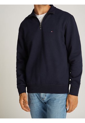 Homme portant un pull bleu marine à fermeture quart de zip avec un petit logo sur la poitrine et un jean bleu clair, debout devant un fond uni.