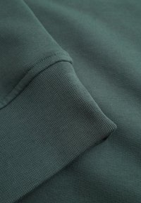 Mørkegrønt sweatshirt-stof med ribbet struktur og et nærbillede af manchet-detaljen, der viser et glat, ensartet væv.
