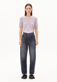 Donna in piedi, di fronte, che indossa un top a maniche corte viola chiaro, jeans neri sbiaditi a vita alta e scarpe nere con lacci e pon pon.