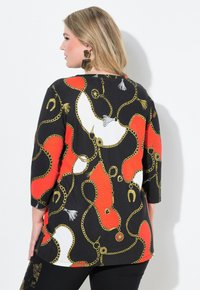 Schwarzes Oberteil mit einem auffälligen Ketten- und Charms-Muster in Rot, Weiß und Gold. Dreiviertelärmel mit lockerer Passform.