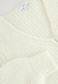 Valkoisessa neulecardiganissa on teksturoitu kuvio, V-pääntie ja yksi kirkas nappi koristuksena. Se on valmistettu pehmeästä, pörröisestä materiaalista.