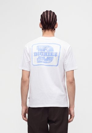 Person mit geflochtenem Haar, die ein weißes T-Shirt mit einem verblassten blauen "Dickies Quality Workwear" Logo auf dem Rücken trägt und mit dem Rücken zum Betrachter steht.