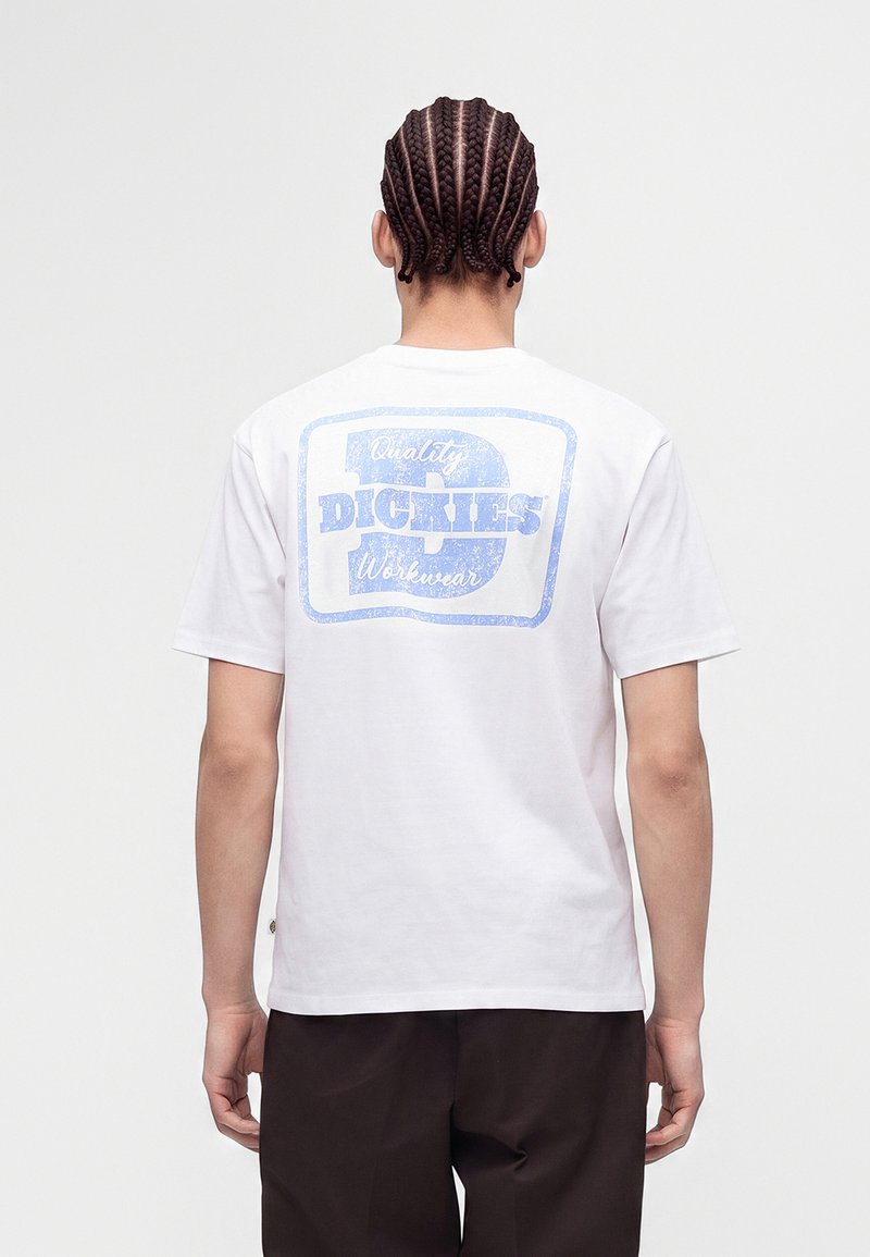 Personne aux cheveux tressés portant un t-shirt blanc avec un logo bleu délavé "Dickies Quality Workwear" dans le dos, de dos.