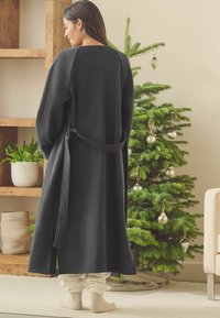 Musta fleece-haalari pitkilla hihoilla ja sidontavyolla, jossa on yksinkertainen muotoilu. Päällä kermaväriset housut ja parina teksturoituja sukkia.