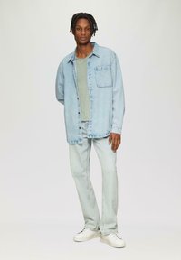 Lichtblauwe denim shirt met knoopsluiting en borstzak, gecombineerd met een lichtgroen t-shirt en lichte spijkerbroek. Witte sneakers zijn te zien.