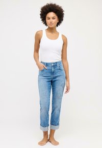 Weißes Tanktop mit blauen, hoch taillierten Jeans, die umgeschlagene Abschlüsse und ein Schmetterlingsstickerei-Detail aufweisen, an einem Modell barfuß vor einem schlichten Hintergrund präsentiert.