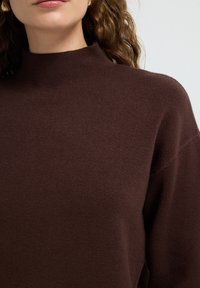 Vrouw in een donkerbruine hoge-nek gebreide trui met golfachtig haar dat gedeeltelijk zichtbaar is tegen een effen achtergrond.