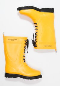 Bottes de pluie en caoutchouc jaunes avec des lacets noirs et une semelle texturée. Présente un logo sur le côté et un accent métallique circulaire près du haut.