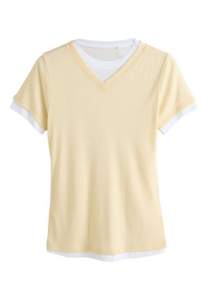 T-shirt jaune clair à manches courtes en V superposé à un top blanc à col rond avec les manches et l’ourlet blancs visibles.