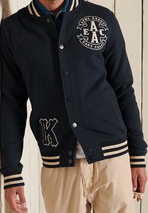 Mand iført navy varsity-jakke med stribede manchetter og kant, mærker med teksten "ATHL ASSOC EAST COAST" og et stort "K" på forsiden.