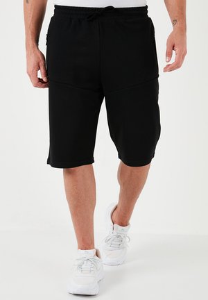Pantaloni sportivi neri con cintura elastica, lacci e tasche laterali. Tessuto liscio e taglio fino al ginocchio, abbinati a sneakers bianche.