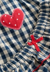 Blau-weißer karierten Stoff mit einer roten gepunkteten Herz-Applikation, roten Nähten und einem kleinen roten Schleifenakzent an der gepufften Rüsche.