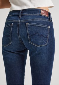 Mörkblå denimjeans med medelhög midja, femficksdesign och en läderlapp på den bakre midjan. Diskret sömnadsdetalj.