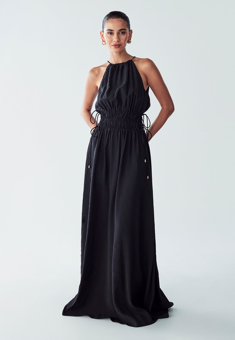 Black halter neck maxi dress with gathered waist, adjustable drawstrings, and a smooth texture. Flowing silhouette to the floor.