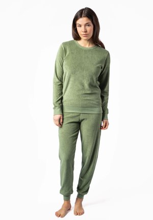 Junge Frau steht barfuß und trägt einen passenden grünen Fleecepullover mit langärmeligem Oberteil und Hose vor weißem Hintergrund.