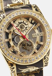 Orologio dorato con cassa trasparente, ghiera decorata con strass e cinturino nero con motivi di drago. Le lancette rosse e nere indicano l'ora.