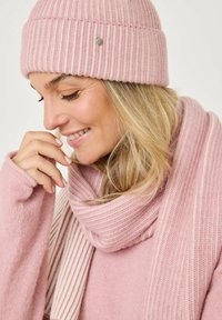 Berretto a maglia a coste rosa chiaro con un accento decorativo in argento; sciarpa morbida abbinata con un motivo a strisce, avvolta attorno a un maglione.