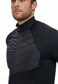 Giacca nera con zip, caratterizzata da un design strutturato, con un pannello imbottito sul petto e un colletto alto. Logo rosso discreto sul petto.
