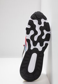 Semelle d’une basket avec une bande de roulement en caoutchouc noire et blanche, accents rouge et bleu visibles sur le côté, positionnée verticalement contre un mur blanc.