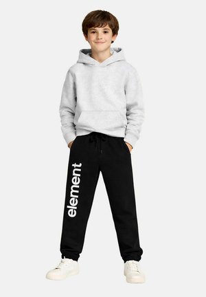 Junge steht mit den Händen in den Taschen, trägt einen hellgrauen Kapuzenpullover, schwarze Jogginghosen mit dem Schriftzug "element" und weiße Turnschuhe vor weißem Hintergrund.