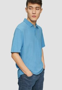 Himmelblaues Polohemd mit Kragen, kurzen Ärmeln und strukturiertem Stoff. Getragen über dunklen Jeans, mit einem einzelnen Knopf am Kragen.