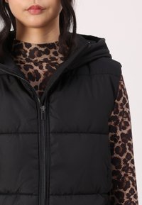 Juodas paminkštintas liemenė su užtrauktuku priekyje ir gobtuvu. Dėvima ant rudos leopardų raštą turinčios palaidinės su aukštu iškirpimu ir ilgomis rankovėmis.