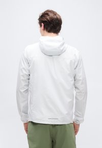 Άνδρας με γκρι ανοιχτό μπουφάν με κουκούλα και χακί πράσινο παντελόνι, πλάτη προς την κάμερα, σε απλό λευκό φόντο.