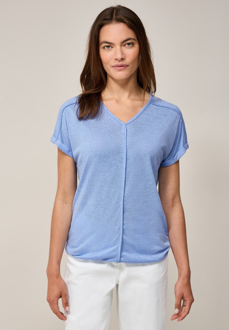 Haut bleu clair à manches courtes et col en V avec des poignets retroussés, présentant une coupe décontractée et une texture subtile. Porté avec un pantalon blanc.
