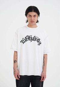 Hvid oversized t-shirt med sort "Ed Hardy" tekst i en stiliseret skrifttype. Har korte ærmer og kontrasterende syninger langs sømmene.
