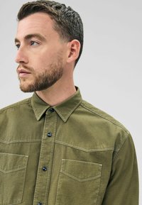 A man with short, styled hair and a beard wears an olive green shirt with two chest pockets, facing slightly left against a gray background.