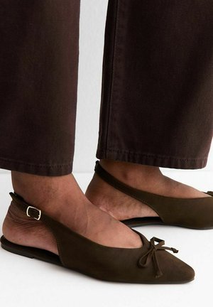 Brązowe baleriny typu slingback z noskiem w szpic i ozdobną kokardą na stopach, zestawione z ciemnobrązowymi spodniami do kostek na jednolitym tle.