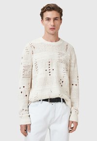 Knit sweater in off-white with openwork patterns and varied textures. Crew neck and long sleeves. Paired with light-colored pants.