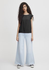 Top nero a maniche corte con dettagli arricciati abbinato a jeans a gamba larga blu chiaro e sneakers in bianco e nero. Sfondo neutro.
