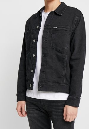 Person iført en sort denimjakke over en hvid skjorte og sorte jeans, stående mod en ensartet lys baggrund.
