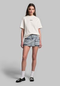 T-shirt bianca in cotone con logo nero, abbinata a una mini gonna di jeans blu chiaro con due tasche frontali, completata con scarpe nere e calze bianche.