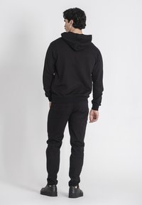 Felpa nera con cappuccio regolabile, polsi ribattuti e vita elastica, abbinata a jeans neri e sneakers nere, vista da dietro.